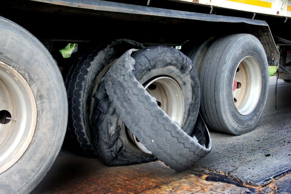 Close-up of a truck with a flat tire, awaiting emergency roadside assistance. The damaged tire is severely worn out, exposing the wheel rim. The truck is stationary on a concrete road, with other intact tires visible in the background.