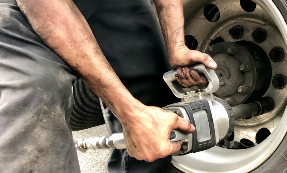 A person with dirty hands and arms operates a power tool to tighten or loosen lug nuts on a large truck wheel. This close-up scene captures the gritty essence of emergency roadside assistance, focusing on the tool and the worker's skilled hands.