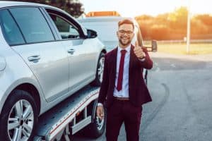 A man in a maroon suit smiles and gives a thumbs up as an emergency truck provides roadside assistance, unloading his car from a tow truck on a sunny day.