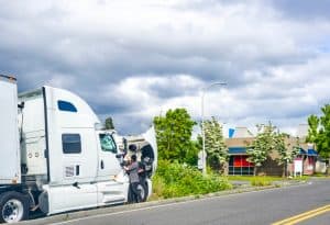 A person examines the open hood of a white semi-truck parked on the side of a road, possibly awaiting emergency truck roadside assistance. The background features a cloudy sky, green trees, and a building with blue and pink accents.
