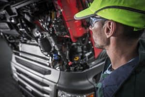 A mechanic wearing a neon yellow cap and safety glasses inspects the engine of a large truck, with the hood open. The focus is on the intricate machinery and components, showcasing his expertise in truck repair.