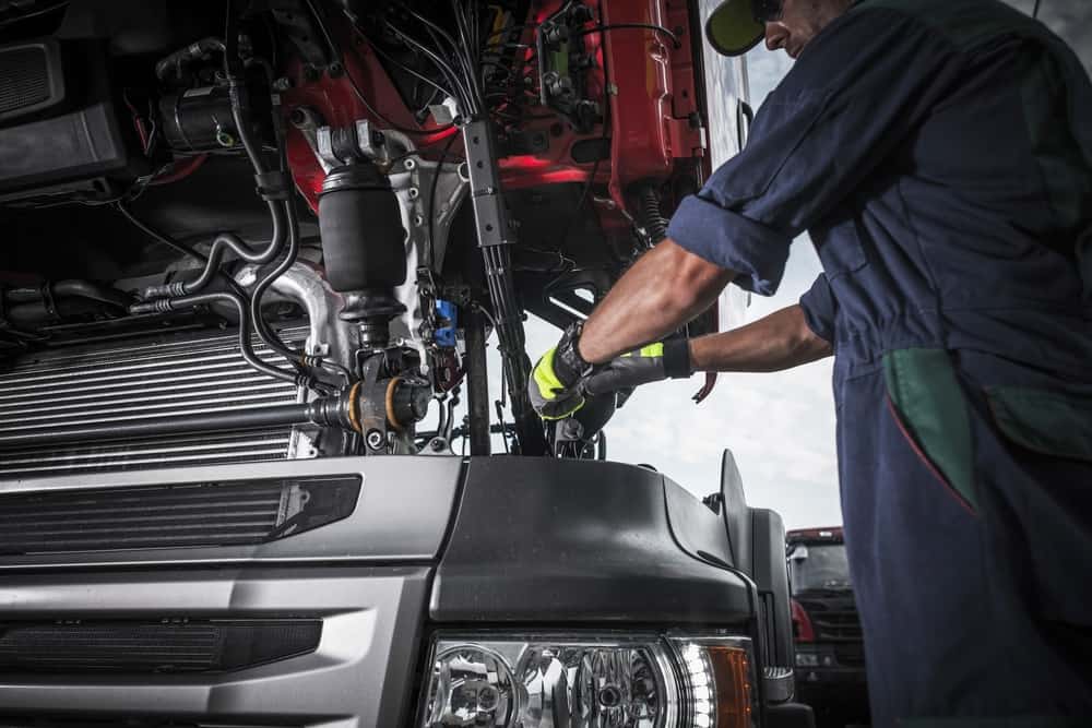 A mechanic in gloves and overalls is inspecting and repairing the engine of a large truck with the hood open. The focus is on intricate components and the mechanic's hands working with tools, showcasing expert truck repair. The silver truck boasts a red cabin interior.