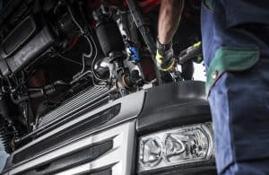 A mechanic skillfully works on a large truck engine, with the hood open for emergency truck roadside assistance. The focus is on the intricate engine components and the mechanic's gloved hands, while the truck's front headlight peeks into view.