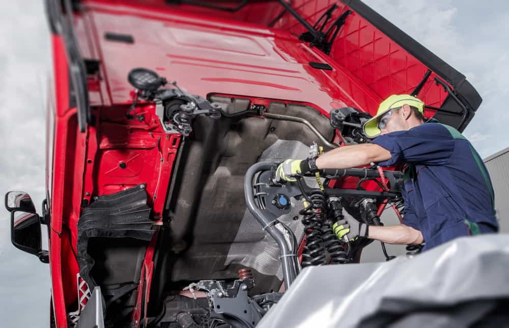 A mechanic in a uniform and cap works intently on the engine of a red truck with the hood raised, providing emergency truck roadside assistance. He uses tools to adjust components against a backdrop of cloudy skies.