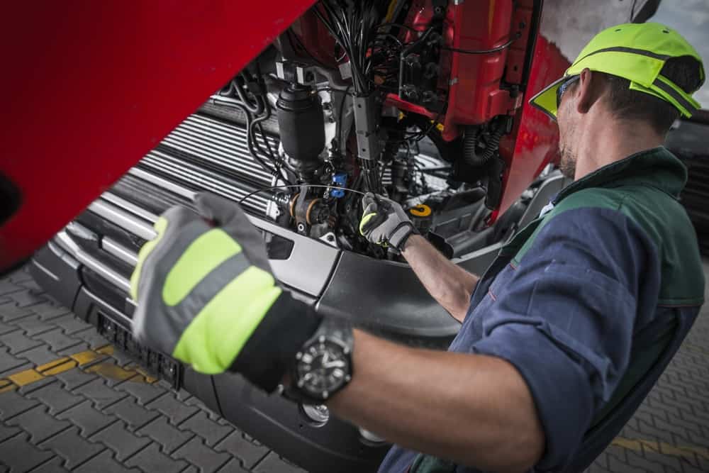 A mechanic in a neon cap and gloves diligently works on the engine of a large vehicle with a red hood open, providing expert roadside assistance. They inspect and adjust parts, standing on a paved surface marked with yellow lines.