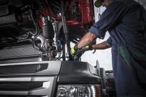 A mechanic in a dark coverall is meticulously working on the engine of a large truck, prepared for emergency roadside assistance. With the hood lifted, focus on intricate parts and wiring is evident. Wearing gloves, they're making precise adjustments or repairs to ensure reliable truck performance.
