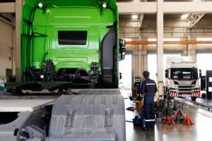 A mechanic in the workshop stands near a large green truck, expertly handling a towing check. In the background, an emergency truck for roadside assistance is lifted on a platform for maintenance. The garage is spacious with high ceilings and industrial equipment.