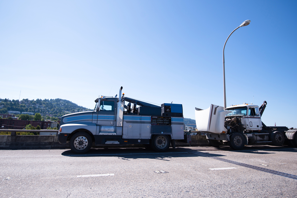 A blue tow truck is parked on a highway under a clear sky, assisting a white semi-truck with its hood raised. A cityscape and green hills are visible in the background.