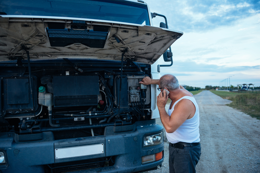 A man in a white tank top stands by a large truck with the hood open, talking on his phone and inspecting the engine on a rural dirt road under a partly cloudy sky.