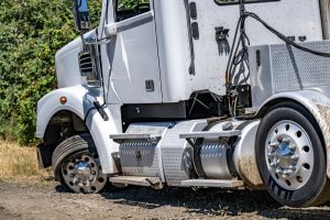 A white semi-truck with a flat front tire is parked on a dirt road near green bushes. The damaged tire is deflated and partially off the rim, and the truck appears to be disabled.