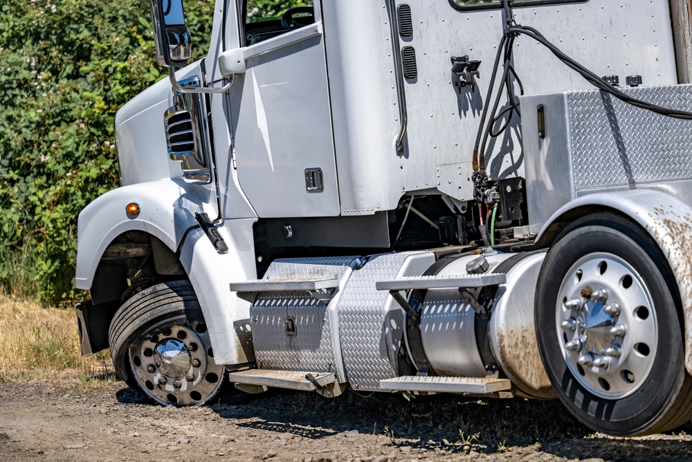 A white semi-truck with a flat front tire is parked on a dirt road near green bushes. The damaged tire is deflated and partially off the rim, and the truck appears to be disabled.