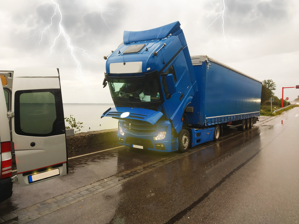 A large blue semi-truck with a severely bent cab is stuck on a wet roadside next to a van, under a cloudy sky with visible lightning in the background.