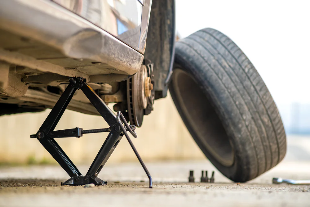 A car is raised on a scissor jack with a removed wheel leaning against the vehicle. Lug nuts and a wrench are on the ground nearby, indicating a tire change in progress.