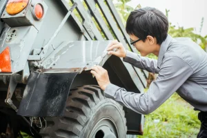 A person wearing glasses and a gray shirt inspects the metal fender of a rugged truck or utility vehicle, examining it closely outdoors in a grassy area.