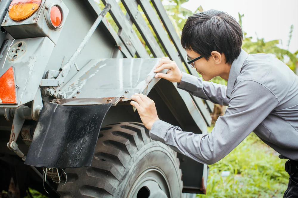 A person wearing glasses and a gray shirt inspects the metal fender of a rugged truck or utility vehicle, examining it closely outdoors in a grassy area.