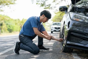 A man kneels beside a car on the roadside, using a tire iron to change the front wheel. Another car is parked in the background, and trees line the road.