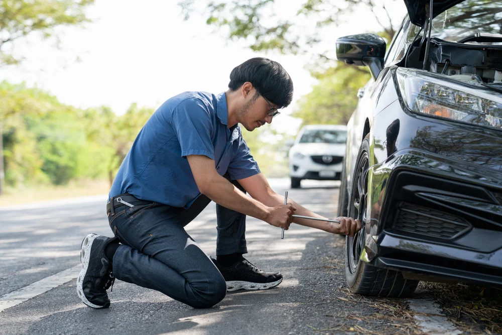 A man kneels beside a car on the roadside, using a tire iron to change the front wheel. Another car is parked in the background, and trees line the road.