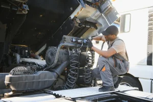 A mechanic wearing a cap and overalls works on the exposed engine of a large truck, using tools and inspecting parts under the raised hood in a well-lit outdoor area.