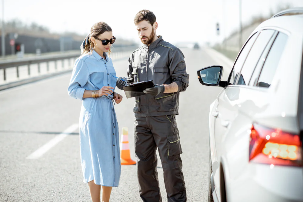 A woman in a blue dress stands by a car on the roadside while a mechanic in work clothes and gloves shows her a clipboard, with traffic cones visible in the background.