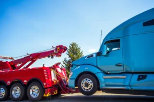 A red tow truck is towing a blue semi-truck during the day. The trucks are parked on a road with a clear blue sky and a tree in the background.