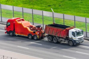 A red tow truck is towing a black and red dump truck on a multi-lane road next to a fence and a grassy area.