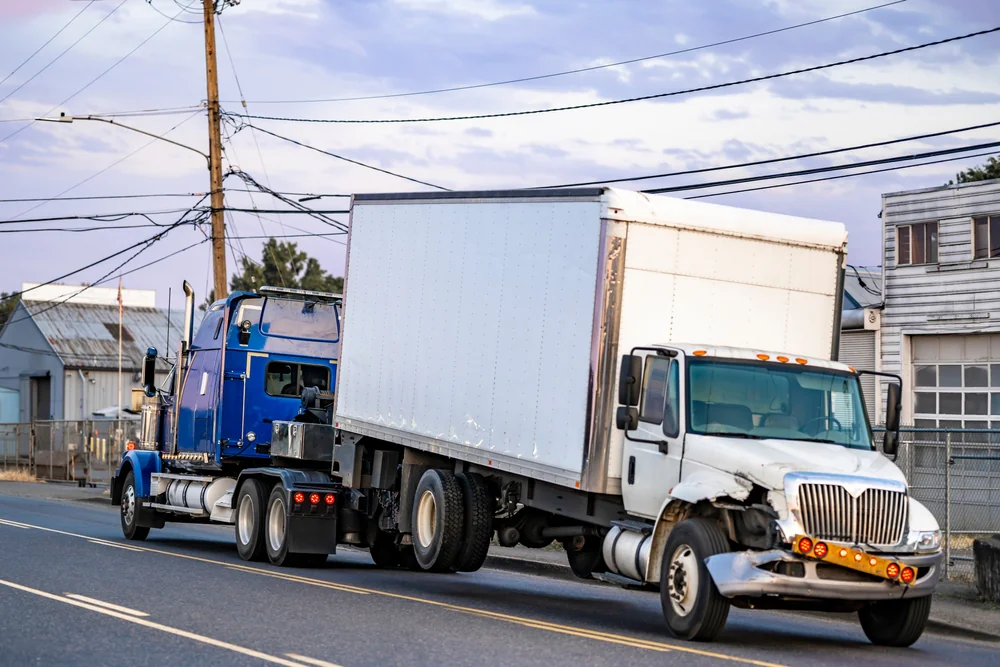 A large tow truck is towing a damaged white box truck with a crumpled front end on a street lined with industrial buildings and power lines.