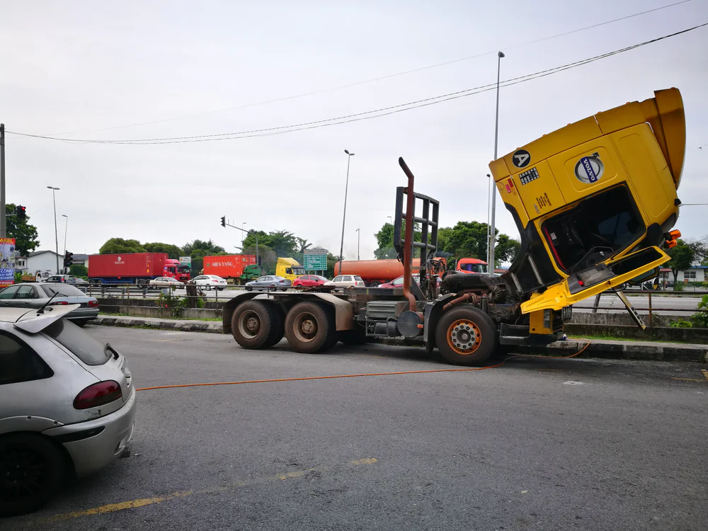 A yellow semi-truck with its cab tilted forward is parked on the side of a road next to a white car. The truck is missing its trailer, and traffic is visible in the background.
