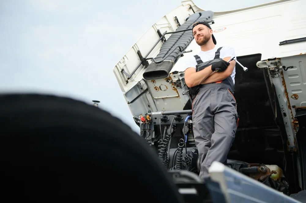 A mechanic in gray overalls and a cap stands with arms crossed in front of a truck with its hood open, looking off to the side. The perspective is from a low angle near a truck tire.