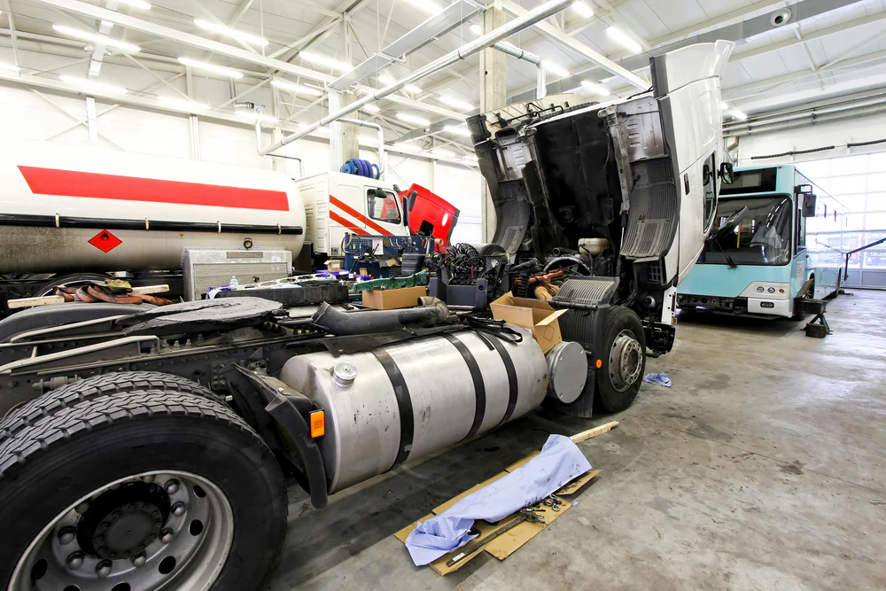 A truck with its cab lifted is being repaired in a spacious, well-lit garage, with tools and parts scattered nearby. Other large vehicles are parked in the background.