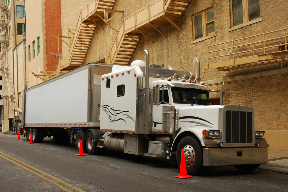 A white semi-truck with a large trailer is parked on a city street next to a brick building, surrounded by orange traffic cones and fire escapes above.