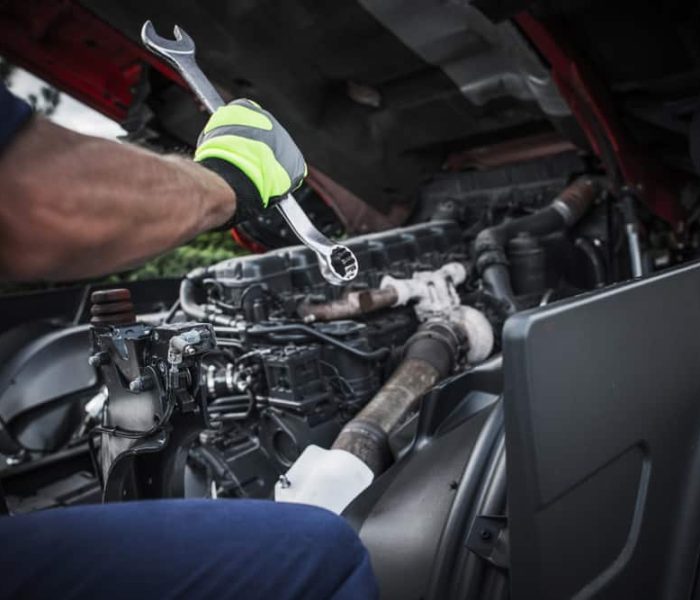 A person wearing safety gloves is providing roadside assistance, working on a vehicle engine with an open hood and holding a wrench. Various engine components and piping are visible in the image.