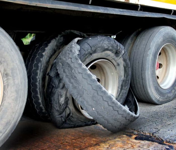 Close-up of a truck with a flat tire, awaiting emergency roadside assistance. The damaged tire is severely worn out, exposing the wheel rim. The truck is stationary on a concrete road, with other intact tires visible in the background.