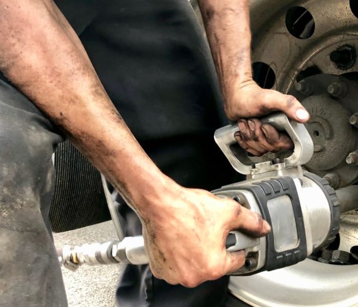 A person with dirty hands and arms operates a power tool to tighten or loosen lug nuts on a large truck wheel. This close-up scene captures the gritty essence of emergency roadside assistance, focusing on the tool and the worker's skilled hands.