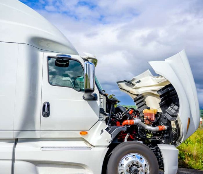 A white semi-truck, its hood raised to reveal engine troubles, awaits roadside assistance. Parked beside a grassy area under a partly cloudy sky, the scene hints at possible truck repair or towing in progress.