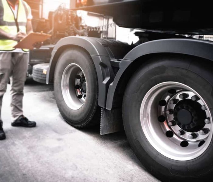 A person wearing a high-visibility vest and holding a clipboard stands next to the large wheels of a truck, emphasizing inspection or roadside assistance work. The image highlights the truck's tires and part of the cab, suggesting readiness for towing or maintenance.