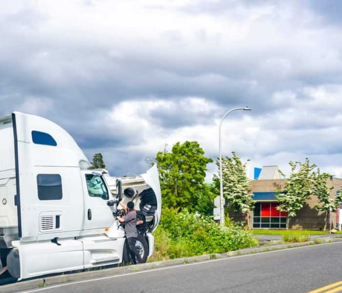 A person examines the open hood of a white semi-truck parked on the side of a road, possibly awaiting emergency truck roadside assistance. The background features a cloudy sky, green trees, and a building with blue and pink accents.